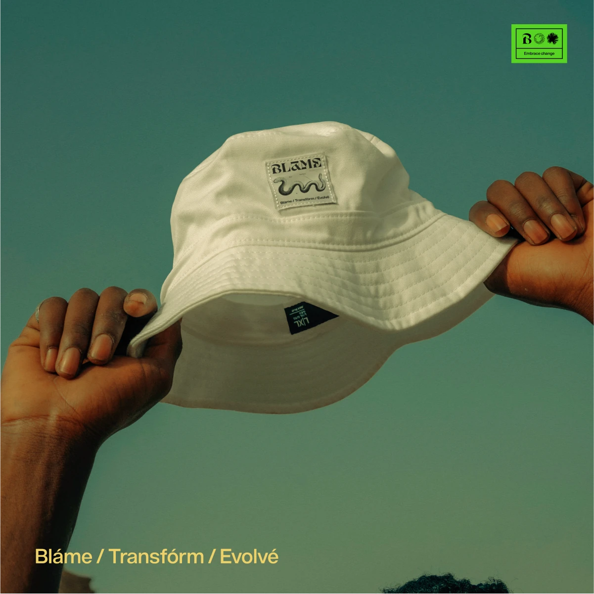 Hands holding up a cream Blame bucket hat with a woven label against a teal sky