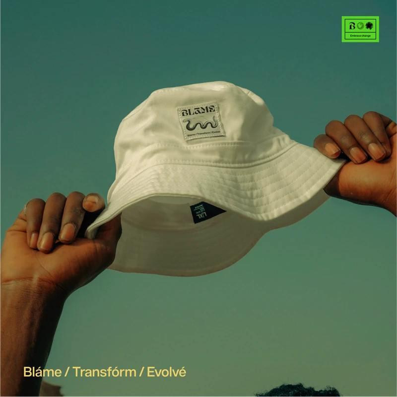 Hands holding up a cream Blame bucket hat with a woven label against a teal sky