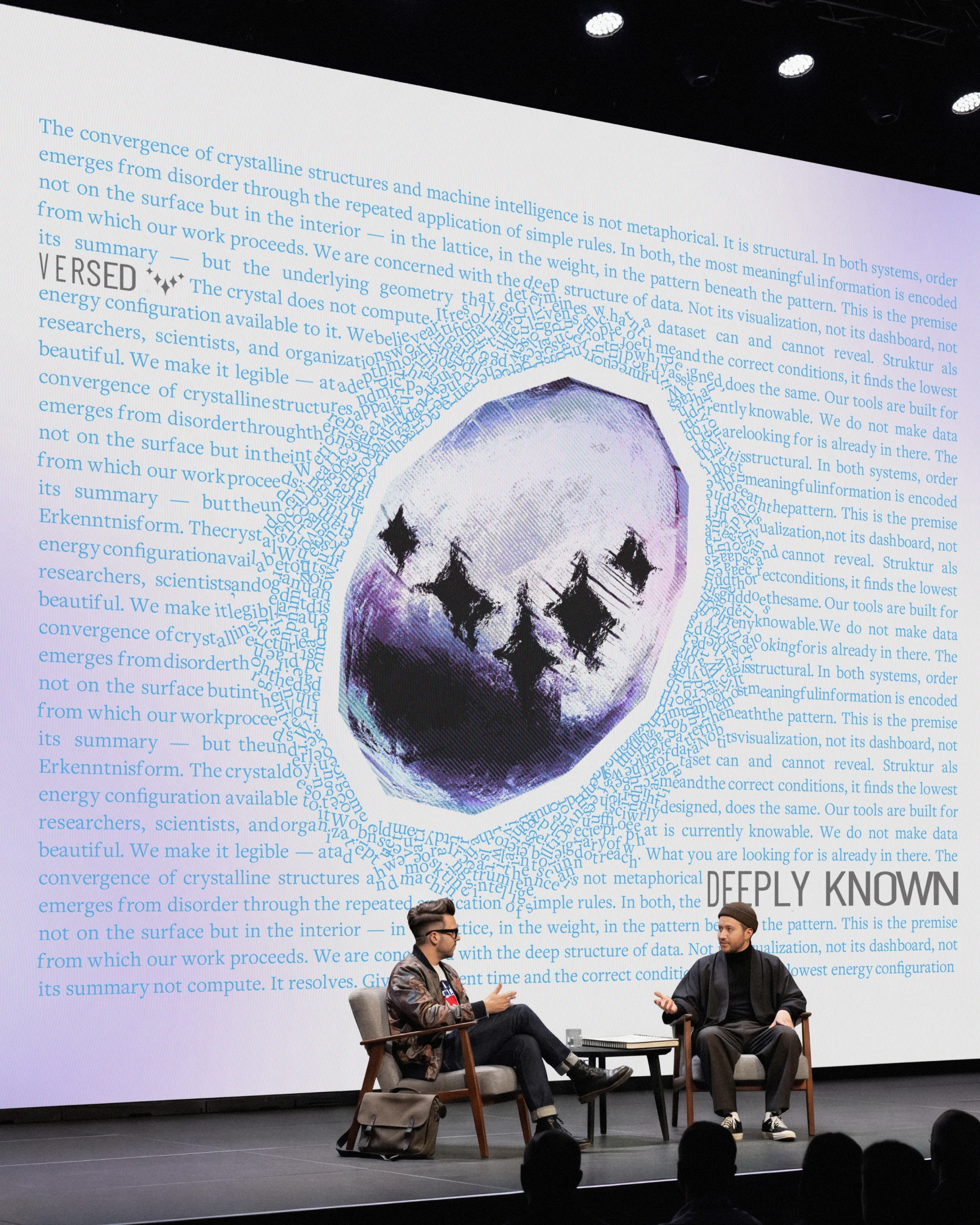 Two people talking on a stage under a Versed screen with a purple crystal ringed by text